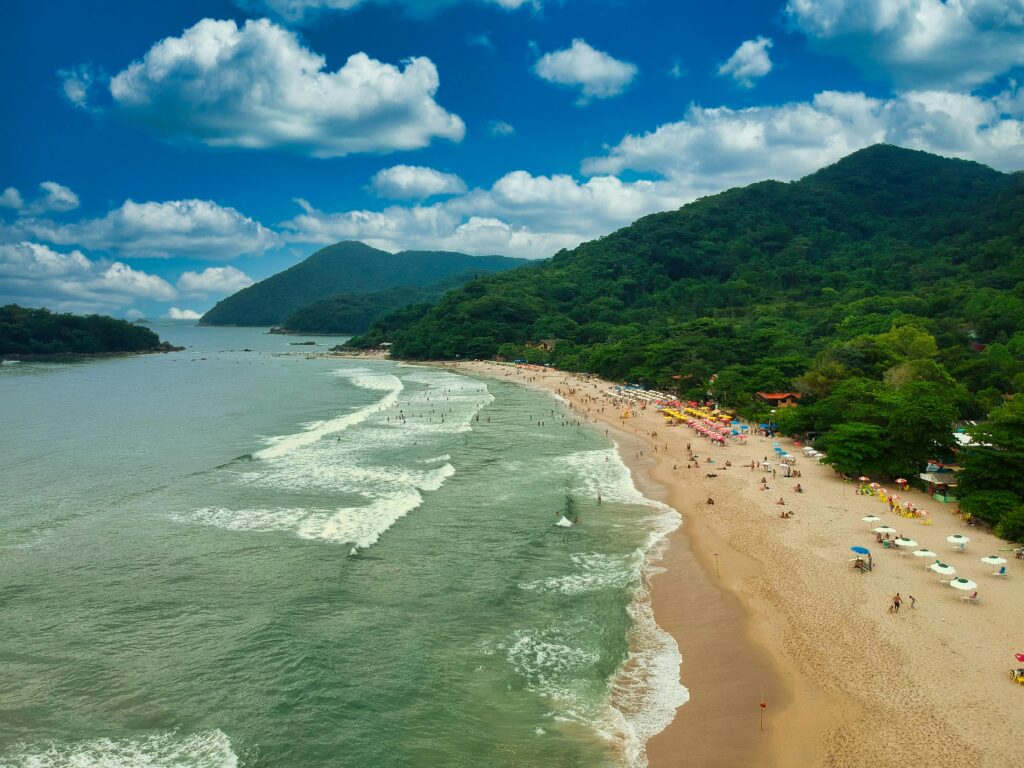Vista aérea da Praia do Curral em Ilhabela, mostrando uma extensa faixa de areia dourada repleta de guarda-sóis coloridos e banhistas, com ondas quebrando no mar esverdeado. Ao fundo, montanhas cobertas por Mata Atlântica se elevam sob um céu azul pontuado por nuvens brancas.