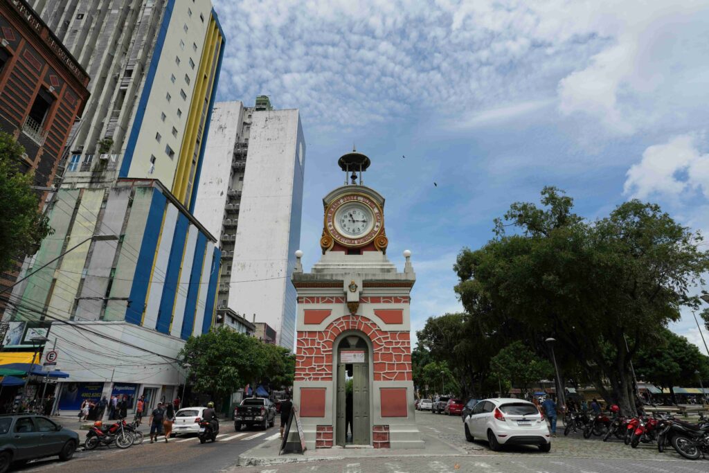 O Relógio Municipal de Manaus em destaque, uma torre histórica no centro urbano da cidade, com edifícios altos, rua movimentada e árvores ao fundo sob um céu parcialmente nublado.