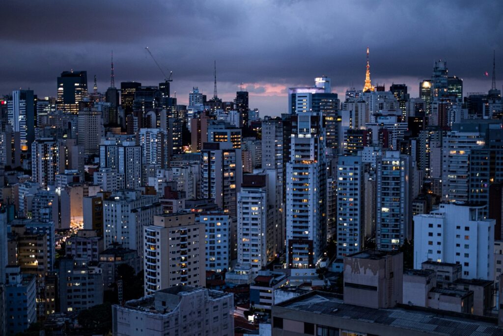 Vista panorâmica da densidade urbana de São Paulo ao anoitecer, com inúmeros arranha-céus iluminados e detalhes de luzes em suas janelas, sob um céu escuro e parcialmente nublado, transmitindo a atmosfera noturna e a grandiosidade da cidade, ideal para ilustrar a vida noturna ou a paisagem urbana de São Paulo.