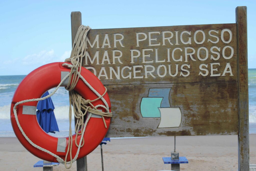 Placa de madeira rústica na areia de uma praia em Porto de Galinhas com os dizeres "MAR PERIGOSO" em português e "DANGEROUS SEA" em inglês, acompanhada de uma boia salva-vidas vermelha, indicando áreas com correntezas ou ondas fortes, com o oceano ao fundo.
