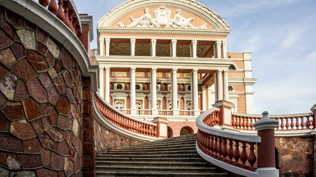 Fachada frontal do Teatro Amazonas em Manaus, exibindo sua arquitetura clássica com colunas, varandas e uma grande escadaria curva em primeiro plano, sob um céu claro.