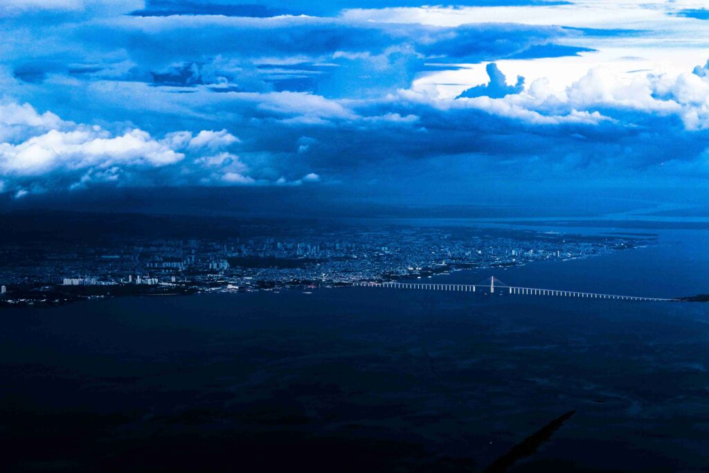 Vista aérea panorâmica da cidade de Manaus e da imponente Ponte Rio Negro sobre o rio escuro, sob um céu dramático e nublado.