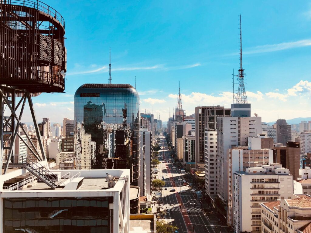 Vista panorâmica aérea da Avenida Paulista em São Paulo, com arranha-céus modernos e antigos alinhados ao longo da via movimentada, sob um céu azul claro, destacando a densidade urbana e a grandiosidade da cidade, ideal para ilustrar roteiros turísticos em São Paulo.