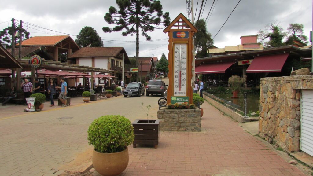 Vista da rua principal de Monte Verde, Minas Gerais, com calçamento de tijolos e construções em estilo europeu, destacando um grande termômetro rústico no centro, araucárias e pessoas caminhando.