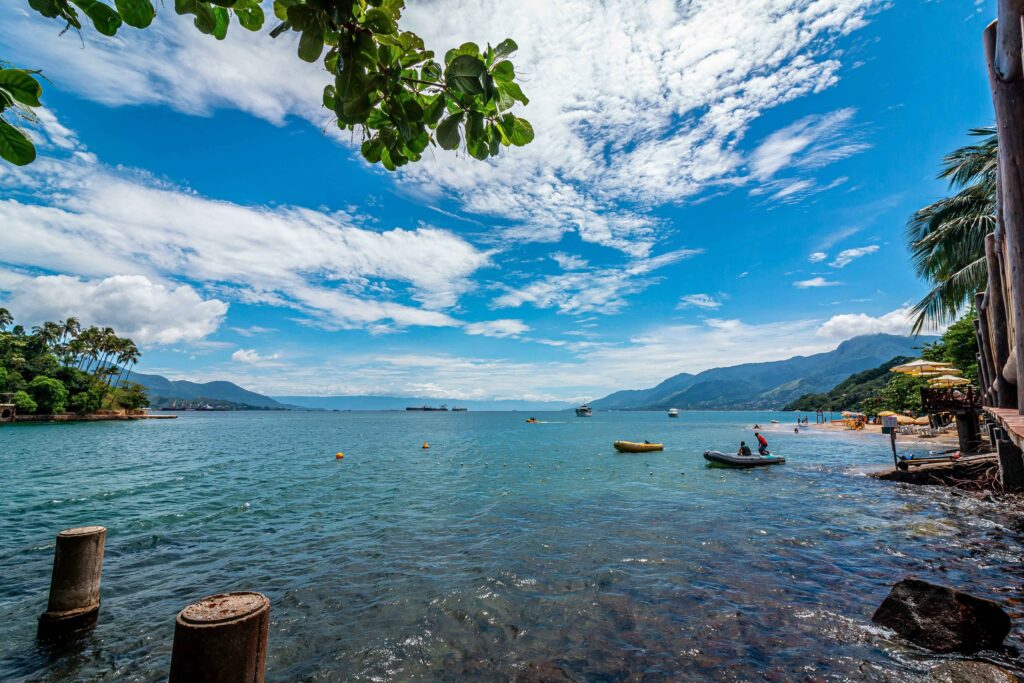 Vista panorâmica de uma baía tranquila em Ilhabela, com águas calmas e transparentes onde flutuam pequenas embarcações. Ao fundo, montanhas verdes se erguem sob um céu azul vibrante com nuvens brancas, enquanto palmeiras e vegetação tropical emolduram a cena, sugerindo a beleza natural da ilha.