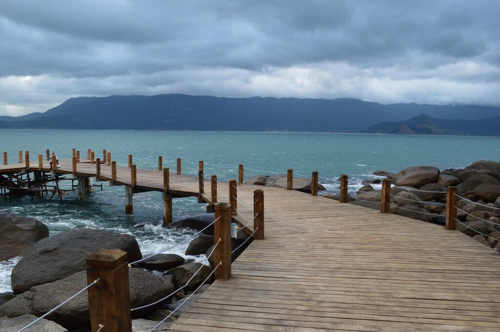 Imagem de um píer de madeira rústico em Ilhabela, estendendo-se sobre rochas e o mar agitado de tom azul-esverdeado sob um céu nublado. Ao fundo, montanhas cobertas por vegetação densa completam a paisagem da ilha.
