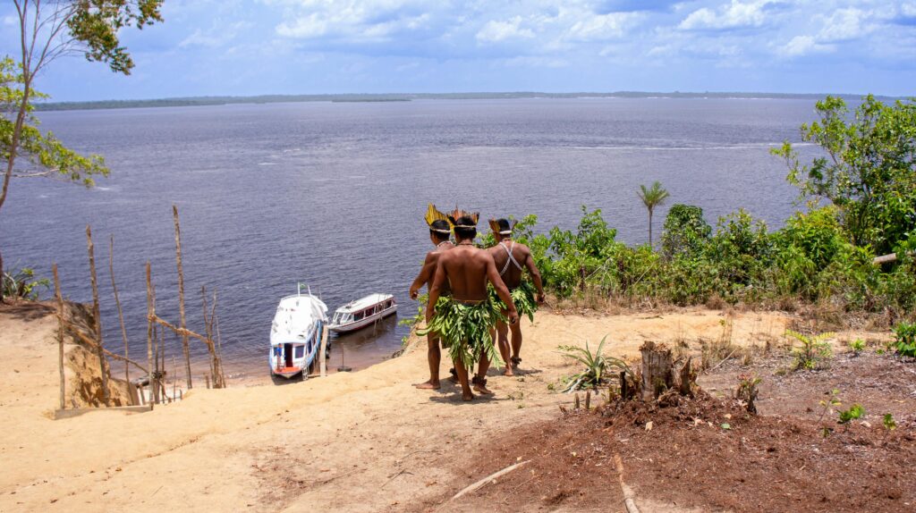 Três pessoas indígenas em trajes tradicionais observam o vasto rio na Amazônia, com barcos na margem arenosa e floresta densa ao redor, representando uma experiência cultural próxima a Manaus.