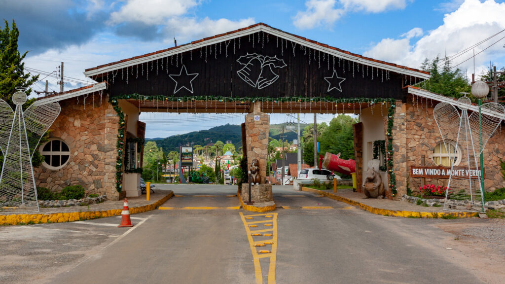 Portão de entrada principal de Monte Verde, Minas Gerais, construído em pedra e madeira escura, decorado com luzes, estrelas e sinos, com a frase "Bem Vindo a Monte Verde!" visível à direita; carros e paisagem montanhosa ao fundo.
