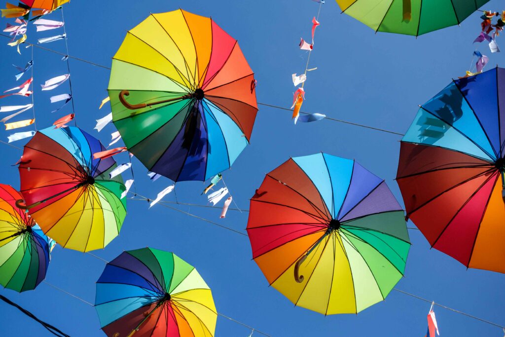 Guarda-chuvas abertos com cores do arco-íris pendurados em fios contra um céu azul claro, criando uma atmosfera alegre e turística em Porto de Galinhas.