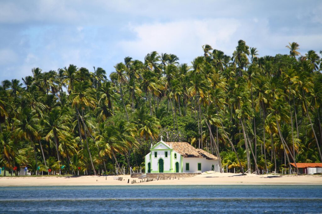 Vista da Igreja de São Benedito (Igrejinha de Porto de Galinhas), uma construção branca com detalhes verdes e telhado colonial, situada na areia da praia, cercada por uma densa floresta de coqueiros e com o mar azul em primeiro plano.