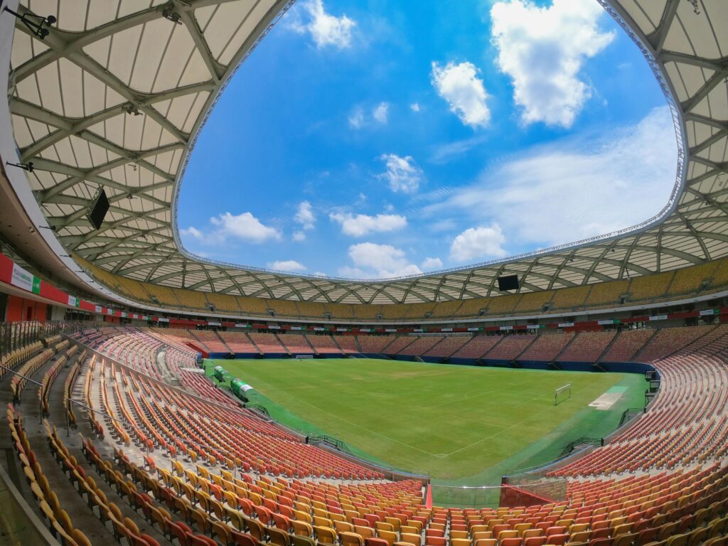 Vista panorâmica do campo e das arquibancadas vazias da Arena da Amazônia em Manaus, com assentos em tons de laranja e amarelo e o céu azul visível através da abertura do teto do estádio.