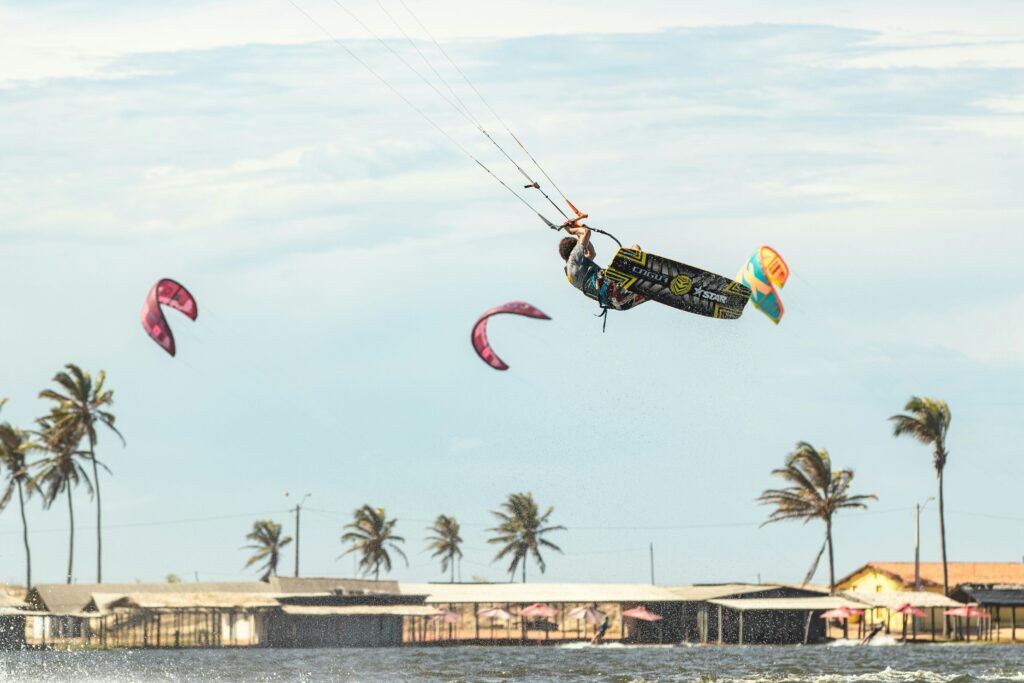 Um praticante de kitesurf realizando um salto impressionante sobre as águas em Jericoacoara, Ceará, com palmeiras e construções rústicas ao fundo, e um céu claro com nuvens esparsas. A imagem destaca a prática de esportes náuticos na região.
