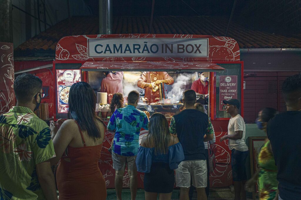 Pessoas aglomeradas em frente a um food truck noturno vermelho, iluminado e com o letreiro "CAMARÃO IN BOX", que oferece refeições rápidas e delivery em Porto de Galinhas, mostrando a movimentação gastronômica da vila.