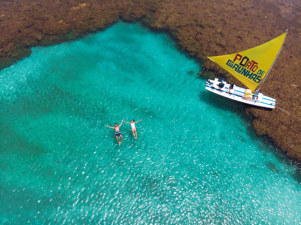 Vista aérea de duas pessoas relaxando e flutuando nas águas cristalinas e azul-turquesa das piscinas naturais de Porto de Galinhas, com recifes de corais escuros ao redor e uma jangada típica com vela amarela "Porto de Galinhas" navegando próximo.