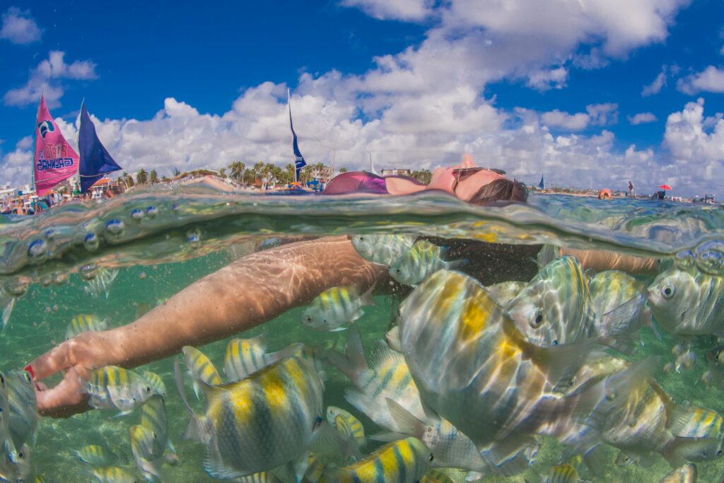 Mulher de óculos de mergulho nadando submersa nas águas transparentes de Porto de Galinhas, cercada por cardumes de peixes coloridos em seu habitat natural, com a superfície da água e o céu azul visíveis na parte superior da imagem.