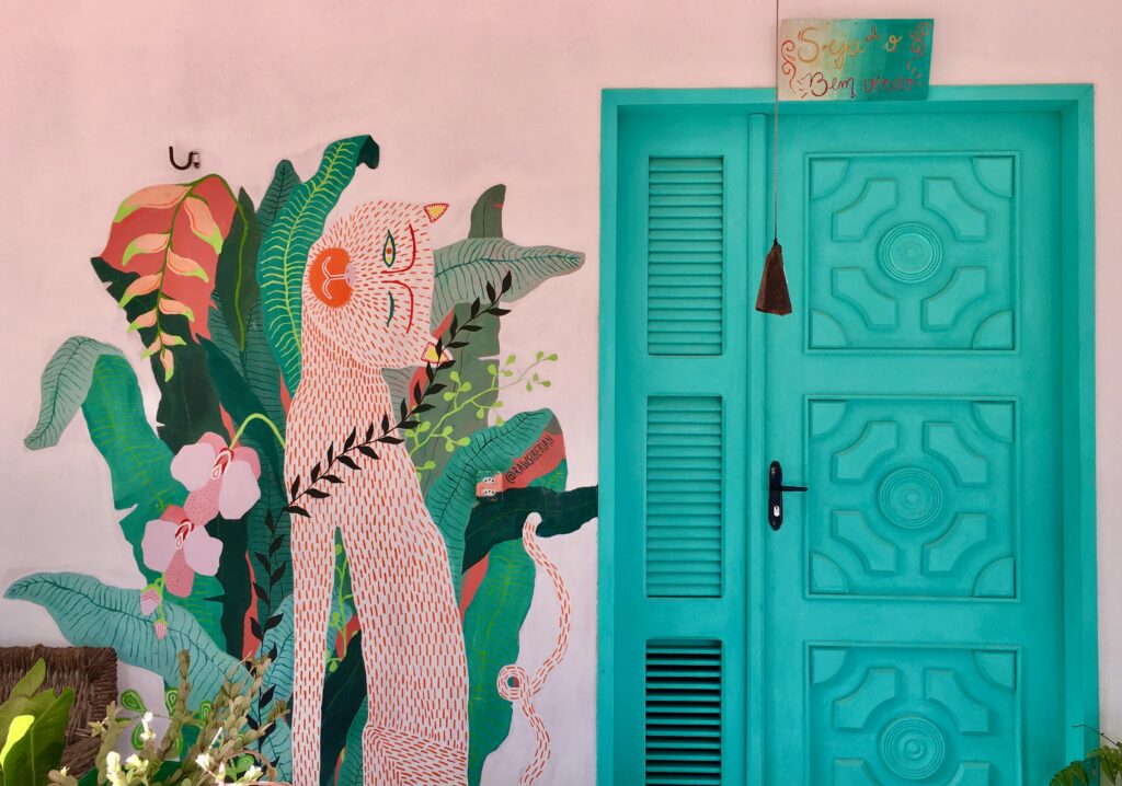Detalhe de uma parede em Jericoacoara com um vibrante mural artístico de um animal estilizado entre folhagens tropicais e flores cor-de-rosa, ao lado de uma porta turquesa com um sino e uma placa de "Seja Bem-vindo" pendurada. A imagem representa a atmosfera acolhedora e artística de Jeri.
