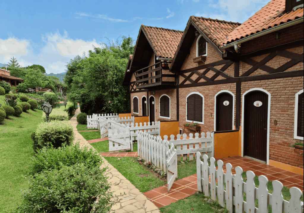 Fachada de chalés em uma pousada de Monte Verde, Minas Gerais, com arquitetura em tijolinho e detalhes em madeira escura, telhados de telha e cercas brancas, cercados por gramado verde e arbustos.