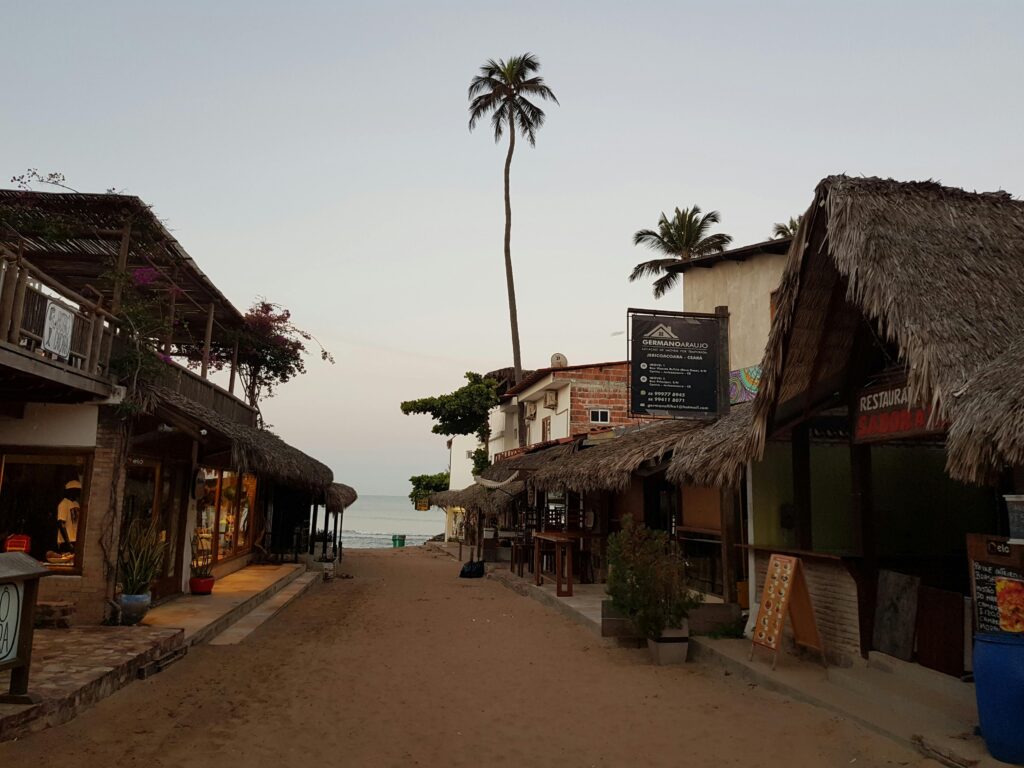 Rua de areia em Jericoacoara, Ceará, ladeada por lojas e restaurantes com telhados de palha e estruturas de madeira rústicas, com palmeiras altas ao fundo e o mar visível no horizonte em um final de tarde.