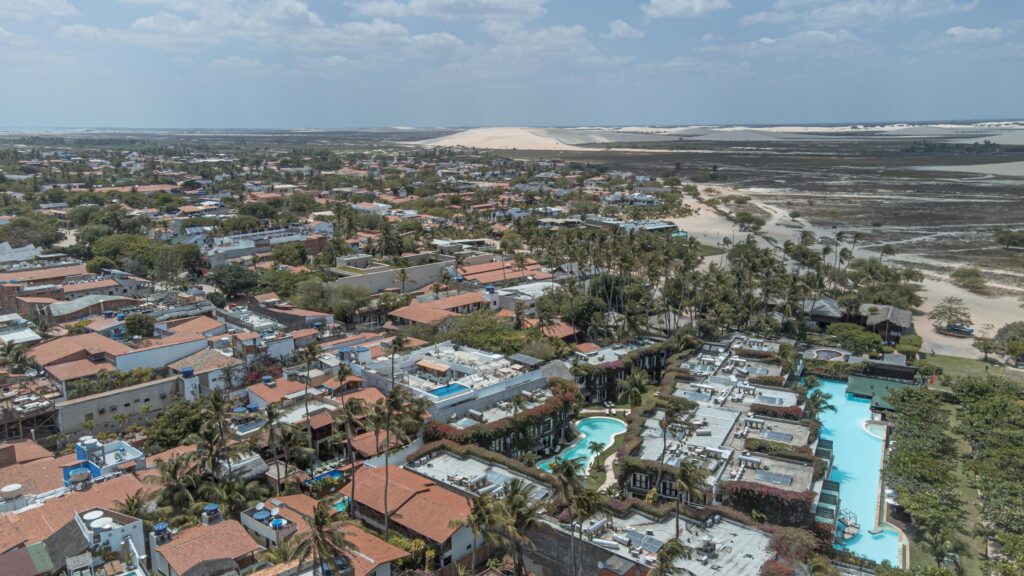 Vista aérea panorâmica da vila de Jericoacoara, Ceará, mostrando telhados alaranjados, coqueiros, piscinas de hotéis e pousadas, com a imponente duna do Pôr do Sol e as lagoas ao fundo, sob um céu azul com algumas nuvens.
