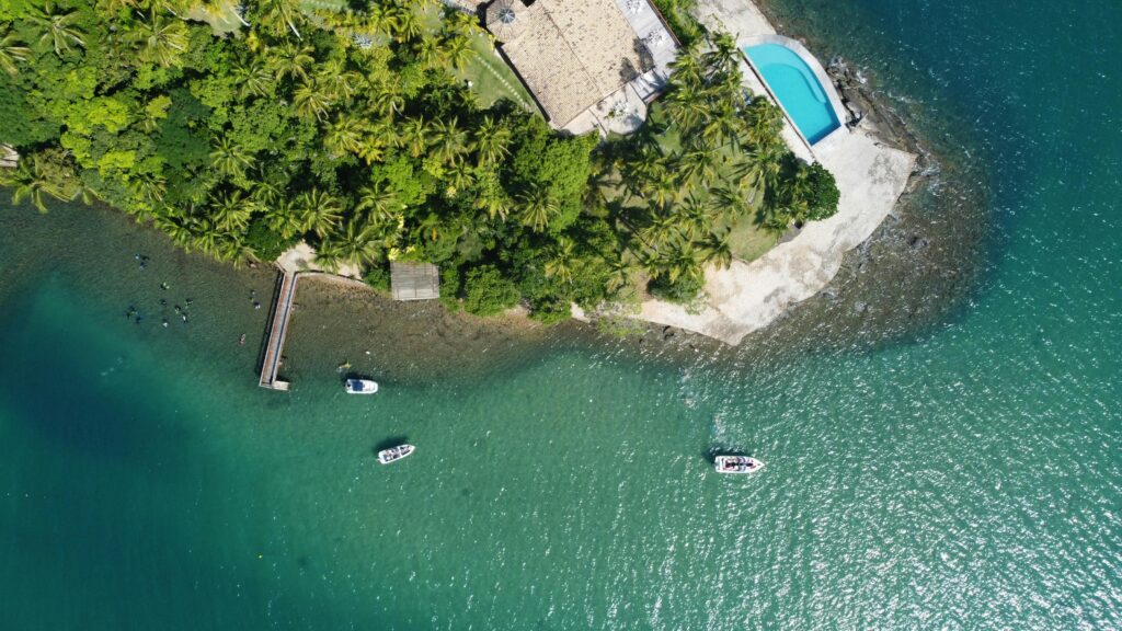 Fotografia aérea de um trecho da costa de Ilhabela, Brasil, mostrando uma densa vegetação tropical que se encontra com águas verde-esmeralda claras. Várias embarcações pequenas estão ancoradas perto da costa e de um pequeno píer de madeira. Uma construção com telhado claro e uma piscina azul turquesa estão situadas na extremidade da faixa de terra coberta por árvores.